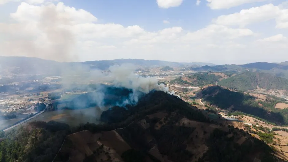 Drone shot capturing smoke rising from a forest fire in a hilly landscape.